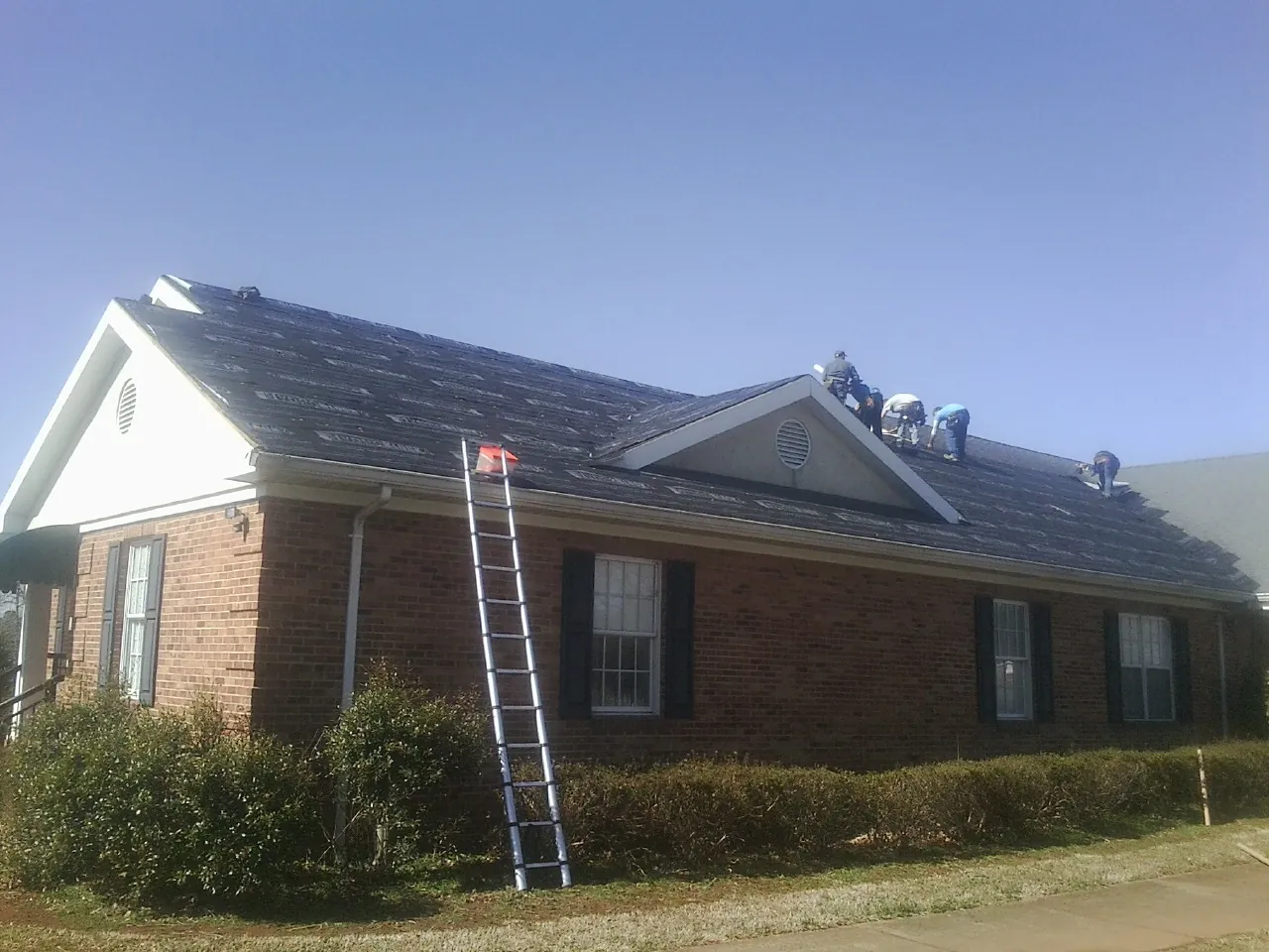 Crew tearing shingles off a brick ranch roof with a ladder set — Emergency Roof Repair work by our crew in Lake Delton