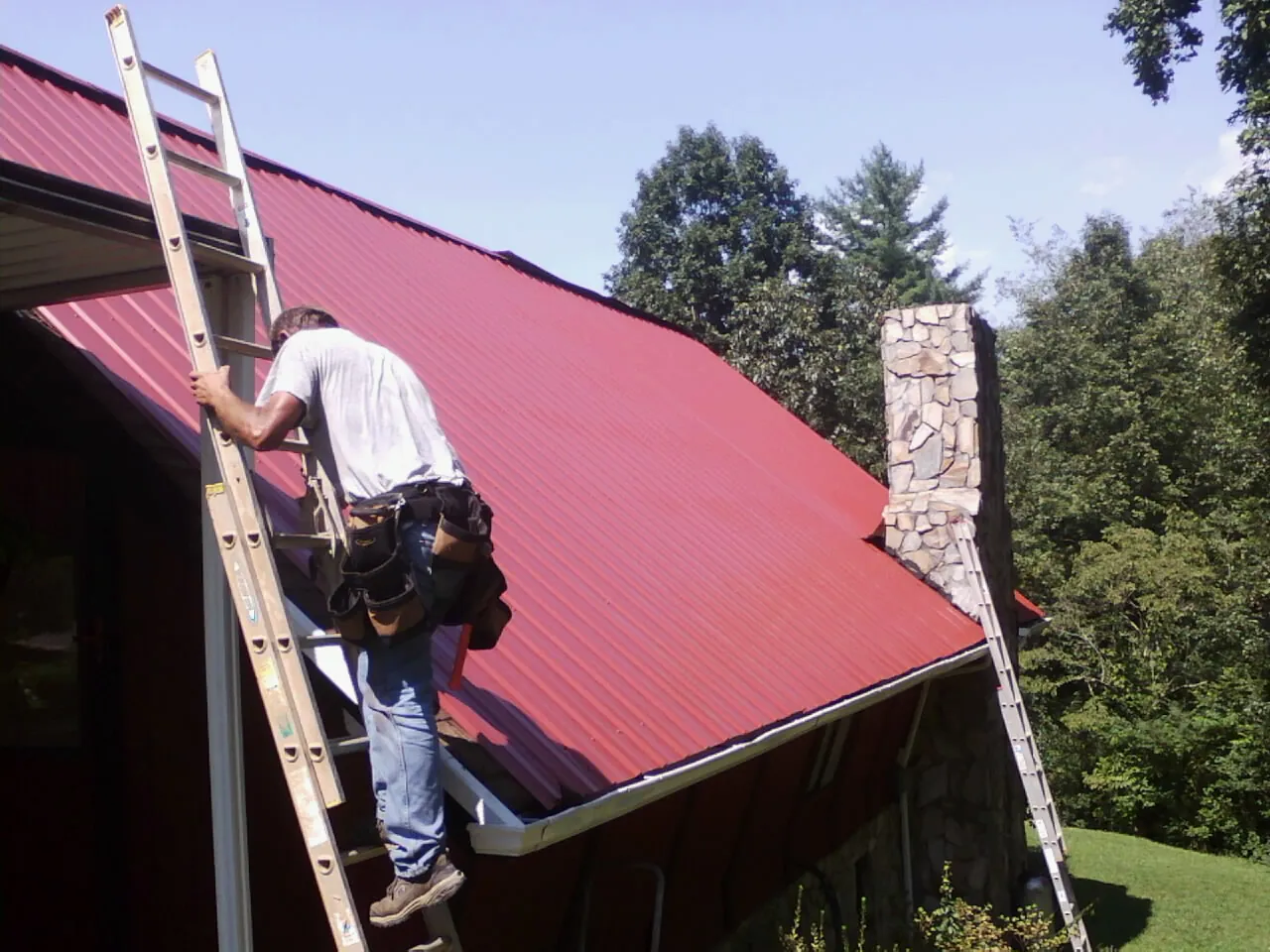 Roofer on a ladder beside a red metal roof and stone chimney — Soffit & Fascia Repair by Roofing Brothers in Lake Delton